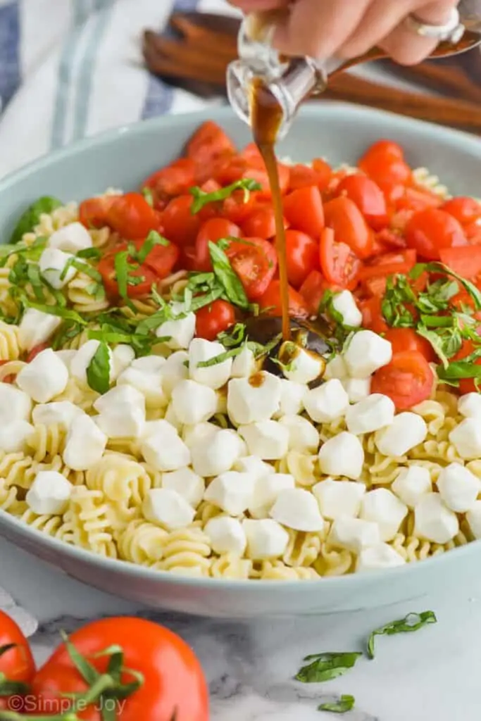 side view of balsamic dressing being poured over pearl mozzarella, pasta, shredded basil and quartered cherry tomatoes