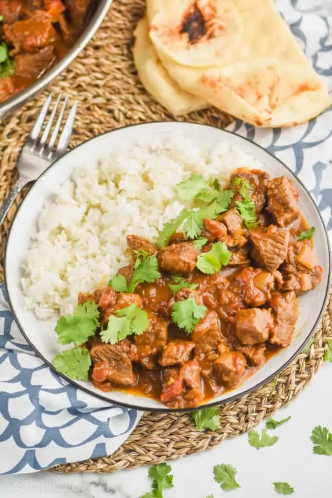 plate with beef curry recipe garnished with fresh cilantro, rice on the other side of the plate and naan in the background