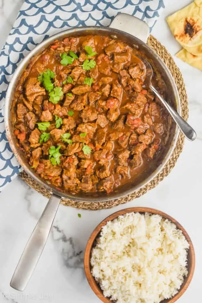 overhead view of a beef curry recipe in a skillet with a bowl of rice next to it and garnished with fresh cilantro