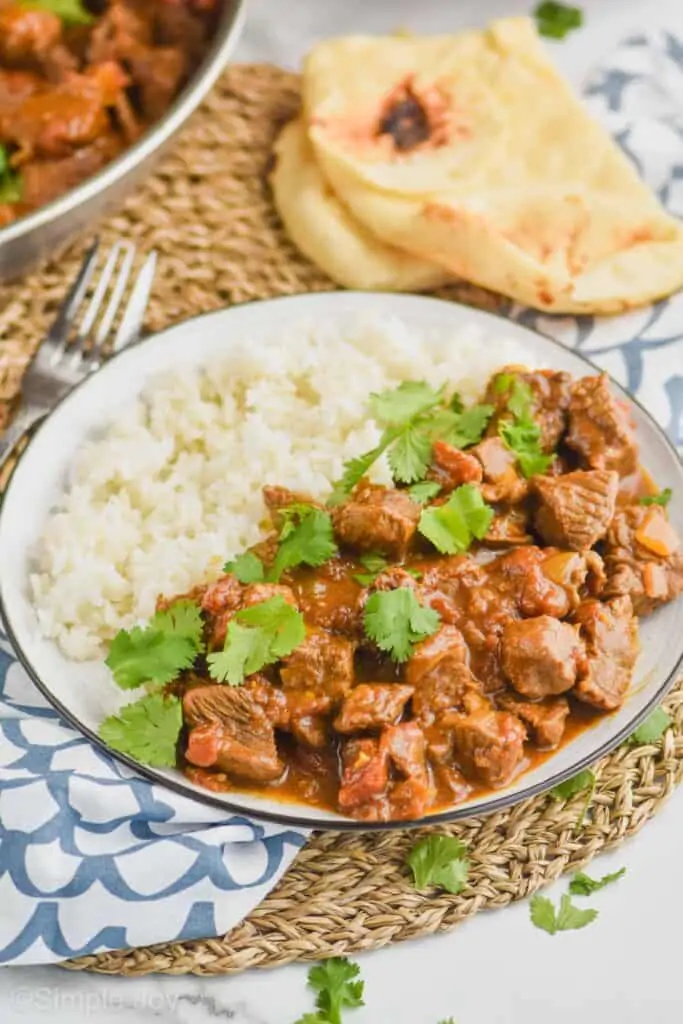a plate with beef curry recipe on one side and and rice on the other side with naan bread in the background