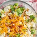 close up of broccoli cauliflower salad in a glass serving bowl with extra shredded cheddar cheese, bacon pieces, sunflower seeds, and diced red onions on top