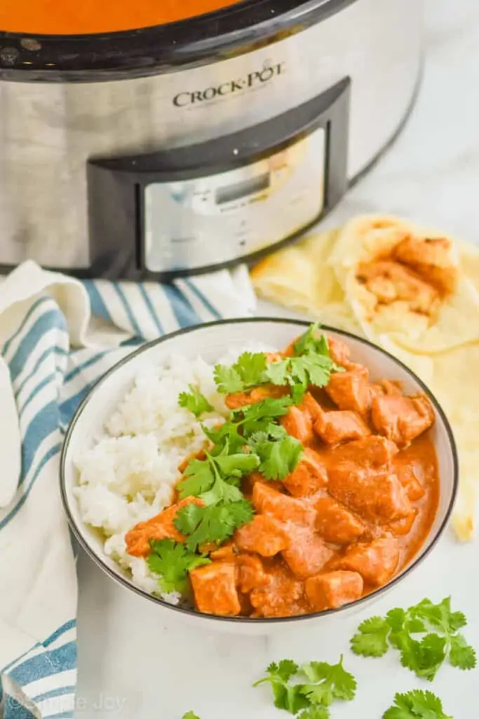 bowl of crockpot butter chicken with naan bread on the side and a slow cooker in the background