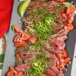 overhead view of cut up up carne asada on a platter with sliced jalapeños, diced tomatoes, sliced limes, and chopped cilantro