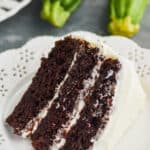piece of three layer chocolate zucchini cake, frosted with cream cheese on a white plate with two zucchinis in the background