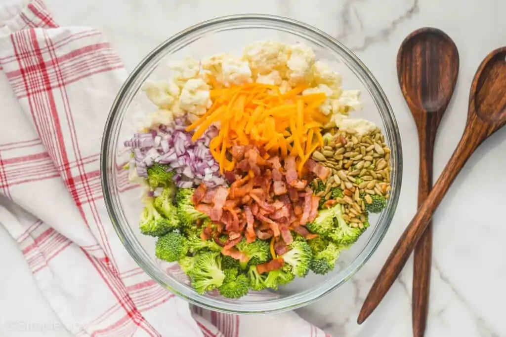 broccoli cauliflower bacon salad ingredients in a clear bowl, each individual ingredient grouped together