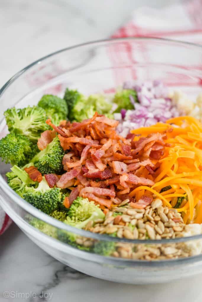 side view of broccoli cauliflower bacon salad ingredients in a clear bowl separated into groups by ingredient