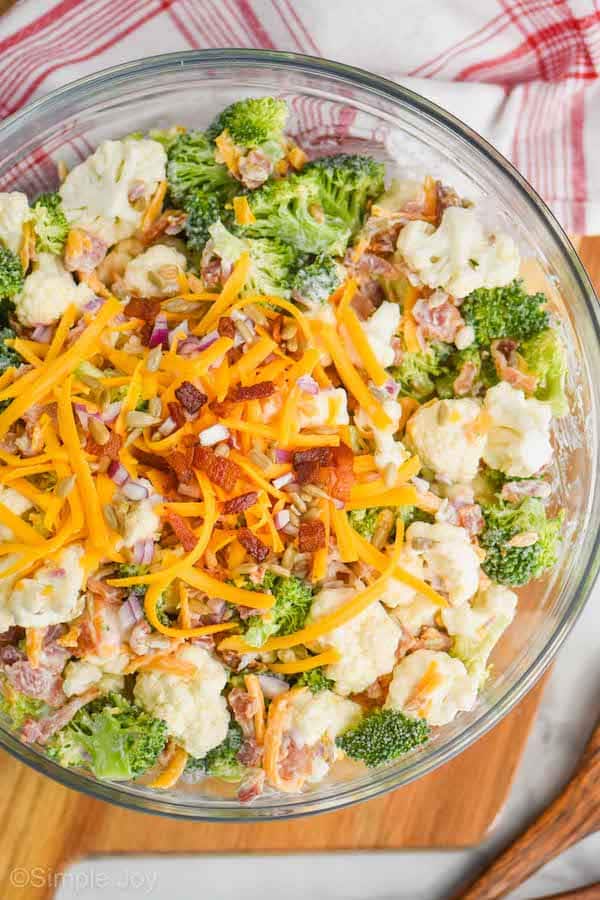 close up of broccoli cauliflower salad in a glass serving bowl with extra shredded cheddar cheese, bacon pieces, sunflower seeds, and diced red onions on top