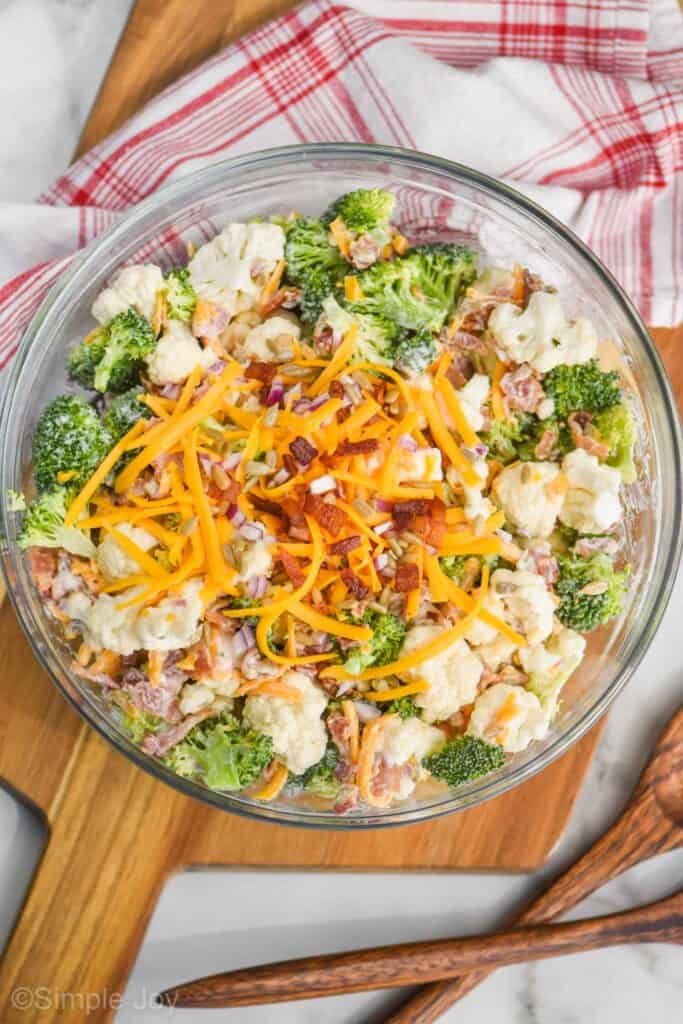 overhead view of broccoli cauliflower salad in a clear serving bowl with extra bacon, cheese, sunflower seeds and red onions on top