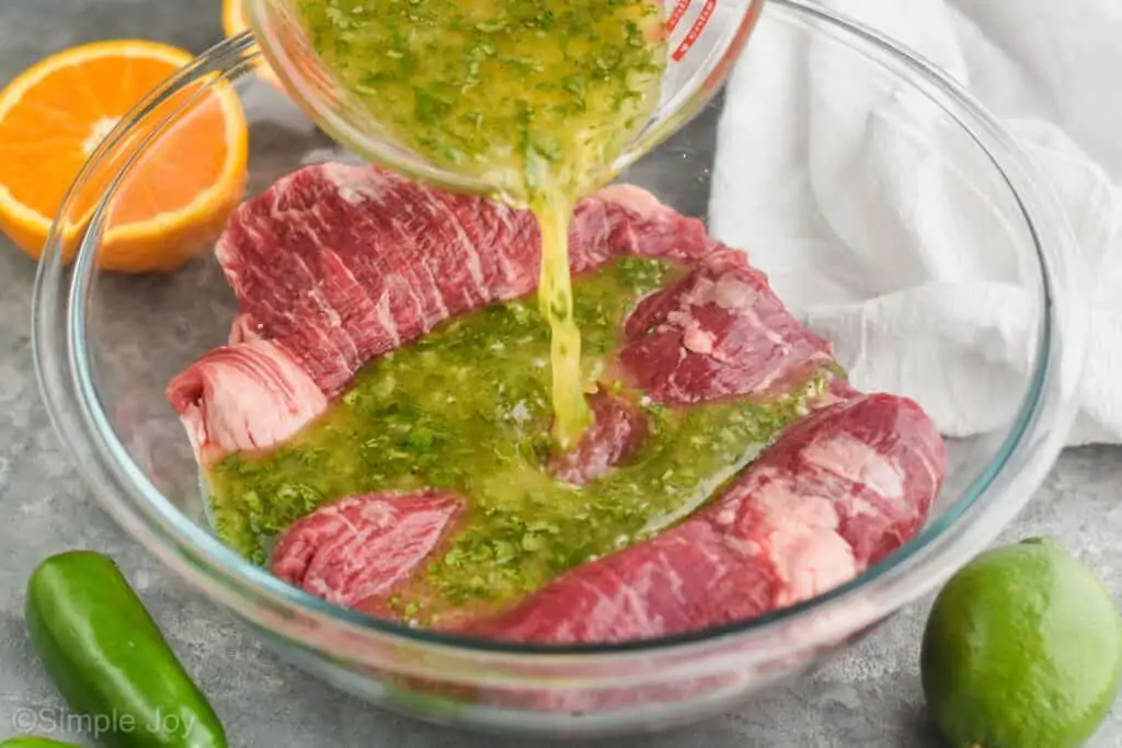 carne asada marinade being poured over skirt steak in a large glass mixing bowl