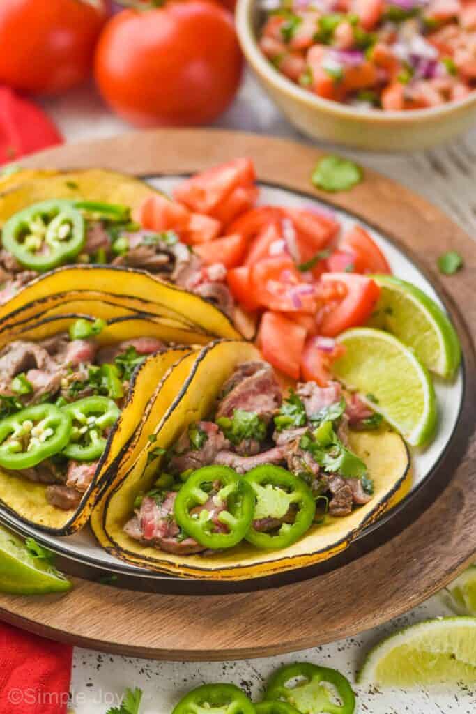 three tacos with carne asada meat in corn tortillas in on a plate garnished with fresh jalapeño and diced tomato and lime wedges on a plate