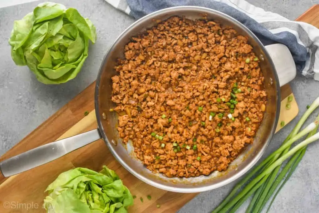 overhead of a pan on a cutting board holding the filling to chicken lettuce wraps with two heads of Bibb lettuce next to it