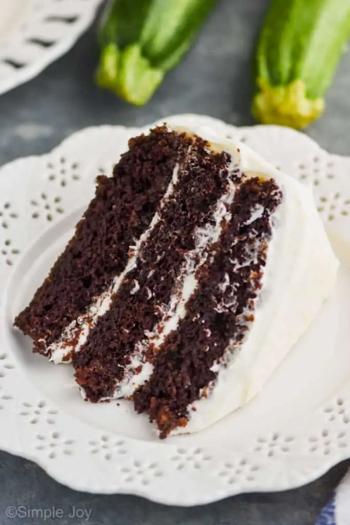 piece of three layer chocolate zucchini cake, frosted with cream cheese on a white plate with two zucchinis in the background