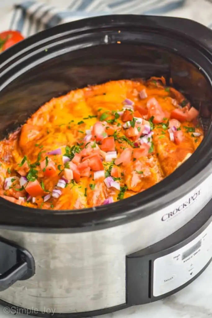 side view of a crock pot holding chicken enchiladas garnished with diced red onion, cilantro, and tomatoes on top