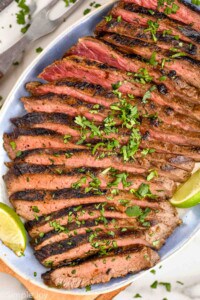 overhead of a grilled flank steak recipe on a platter with cilantro and lime wedges