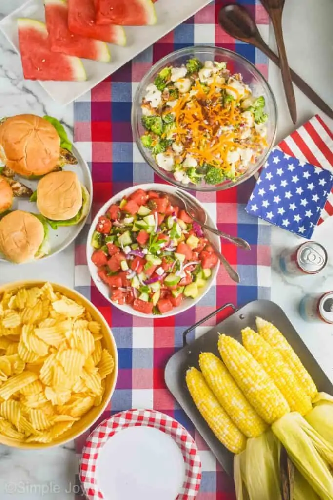 overhead view of a table with food for Fourth of July on a red white and blue checked table runner: watermelon, two salads, burgers, chips, and grilled corn