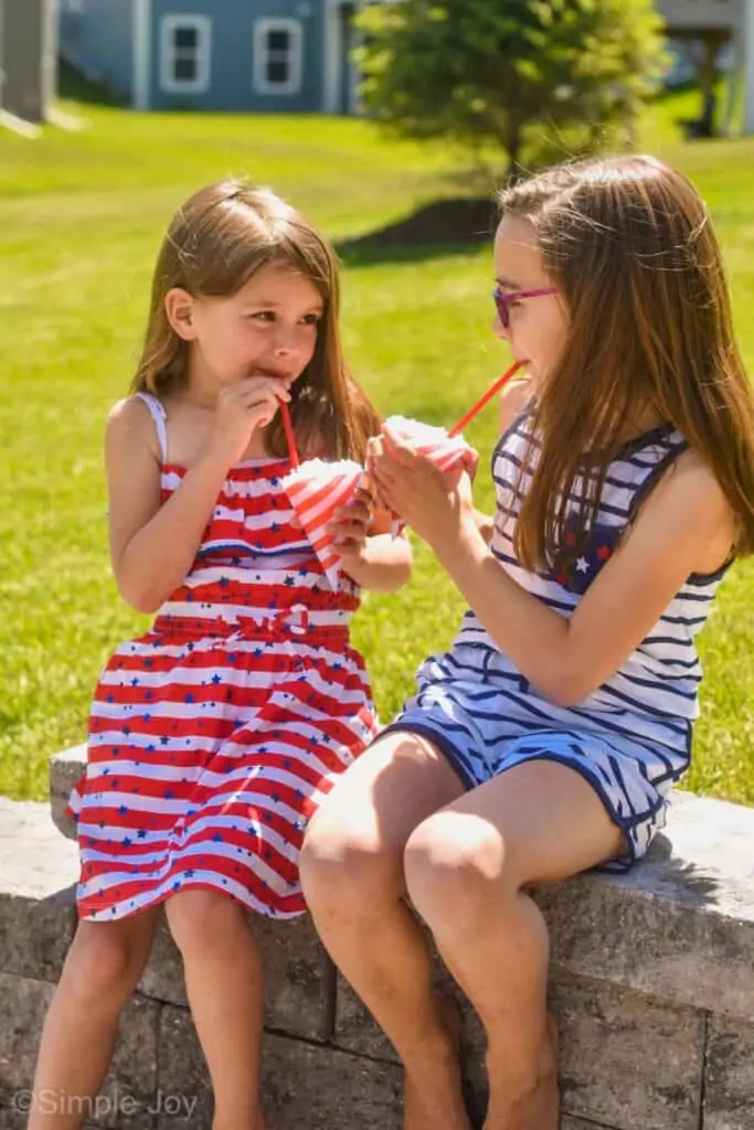 two little girls sitting on a brick wall eating snow cones with straws
