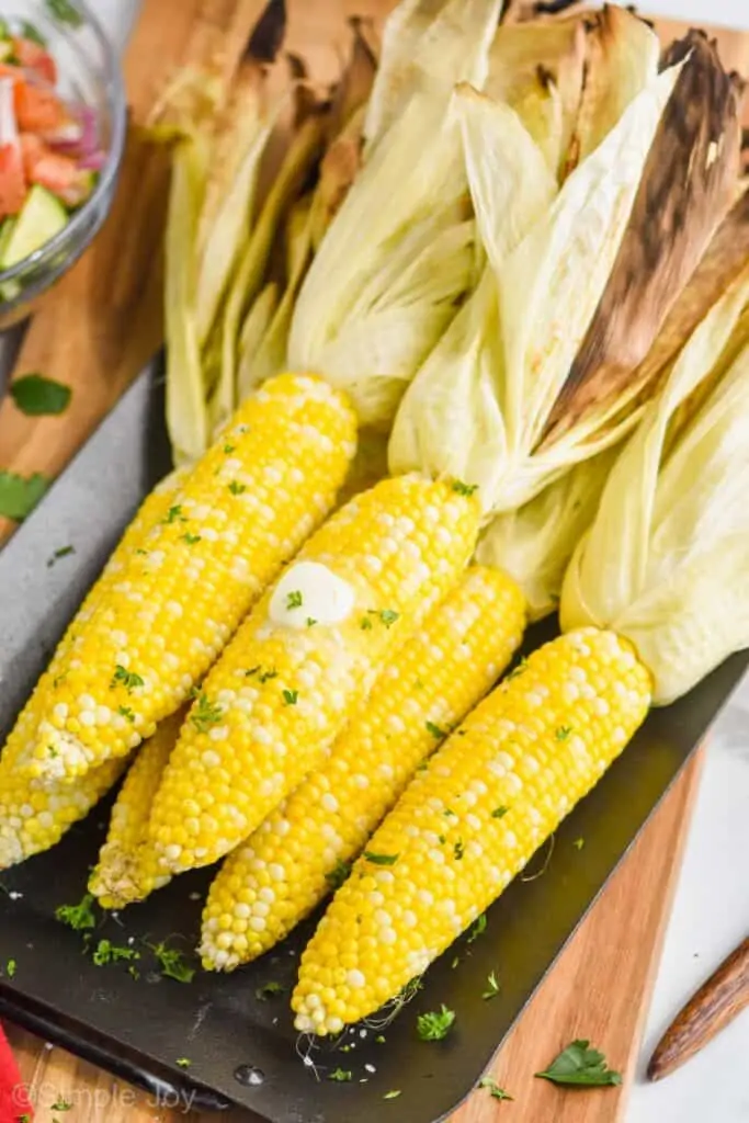 pulled back view of grilled corn on the cob with the husks pulled back that has butter on it and fresh parsley on a platter
