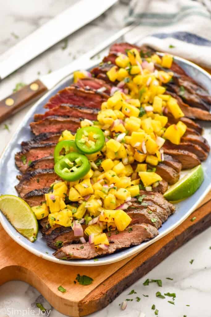 grilled flank steak on a platter, topped with grilled pineapple salsa jalapeno slices and lime wedges