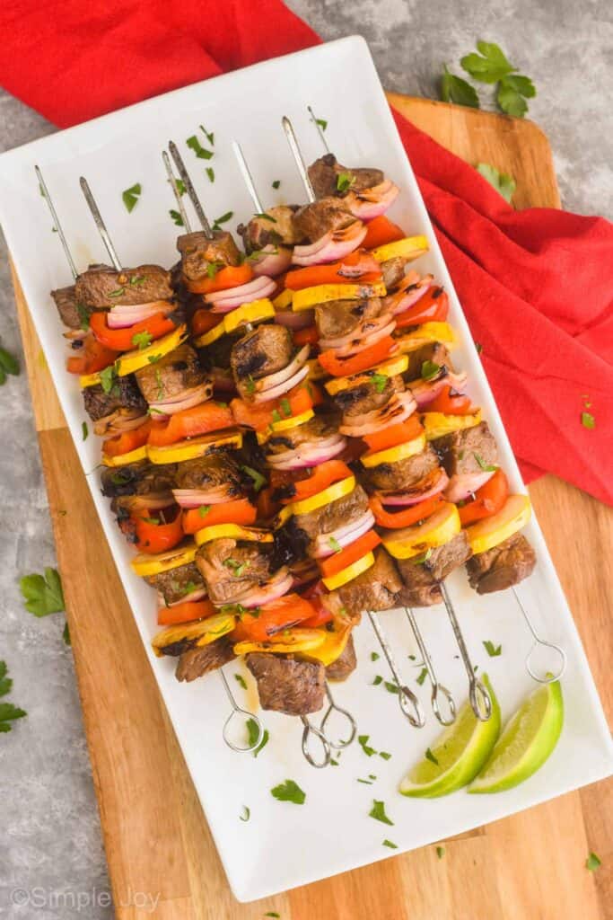 a pile of steak kabobs made with metal skewers on a white rectangular plate, garnished with fresh parsley and with two lime wedges next to them