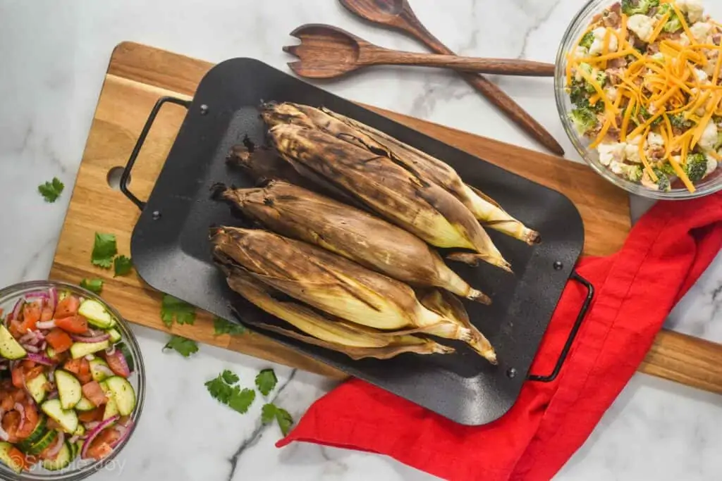 overhead view of grilled corn with the husks still on on a platter on a cutting board