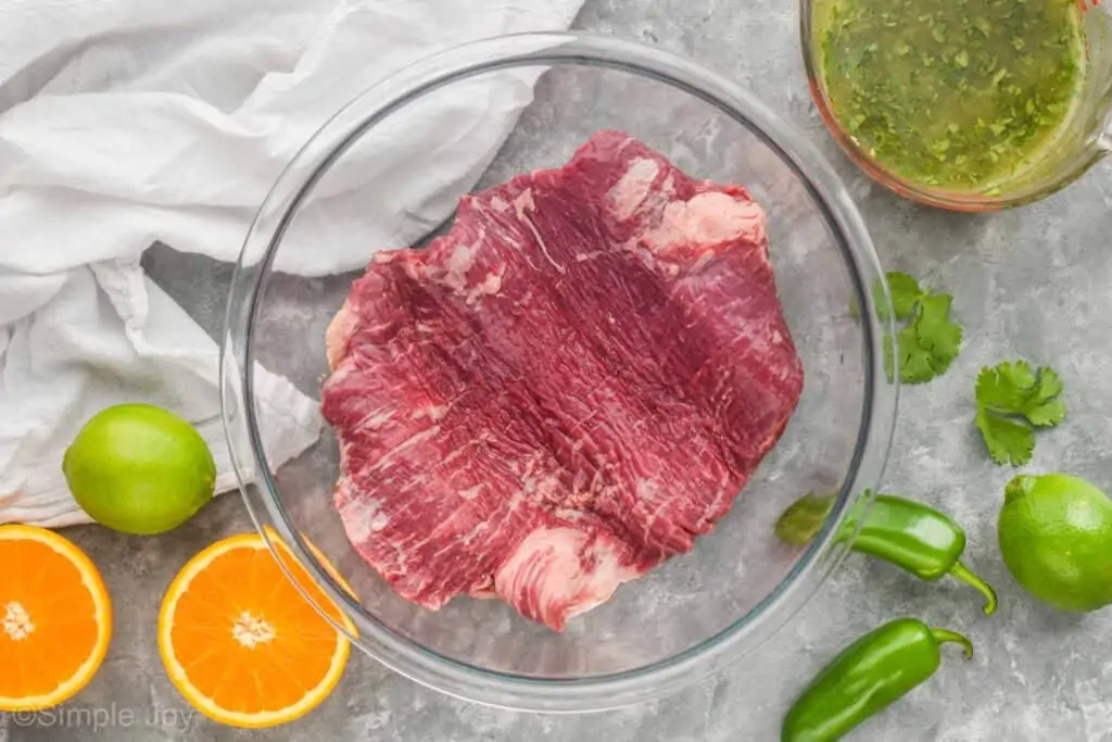 overhead view of a flank steak in a large glass mixing bowl surrounded by an orange, limes, jalapeños, and a marinade