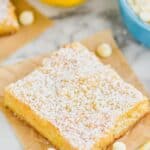 one lemon magic bar topped with powdered sugar on a square of parchment paper with white chocolate chips and lemons around - 6