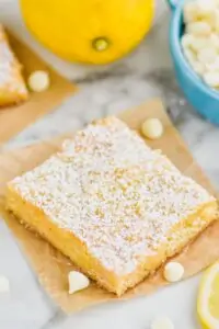 one lemon magic bar topped with powdered sugar on a square of parchment paper with white chocolate chips and lemons around