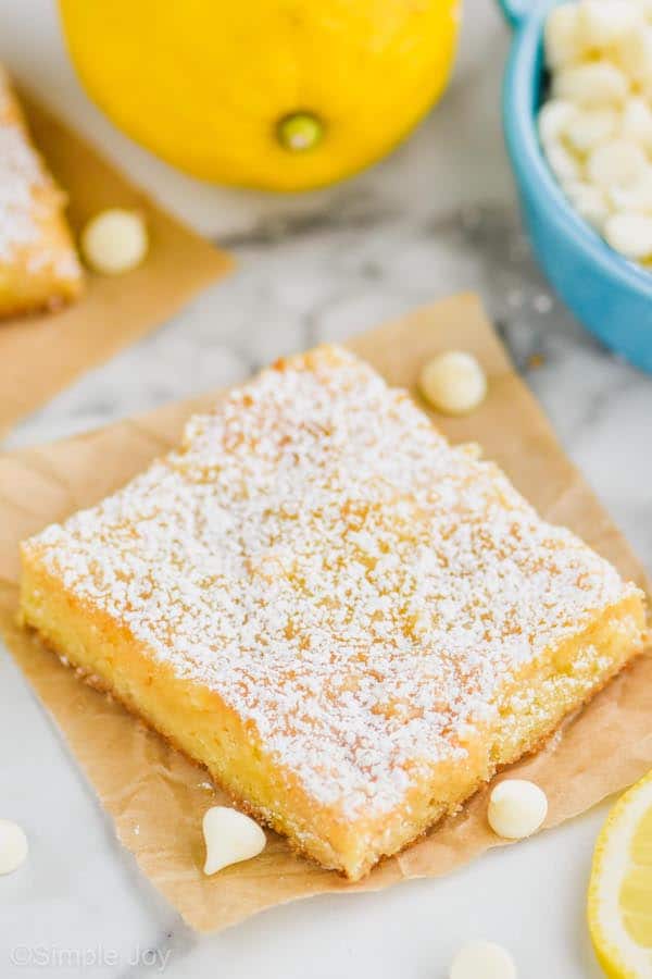 one lemon magic bar topped with powdered sugar on a square of parchment paper with white chocolate chips and lemons around