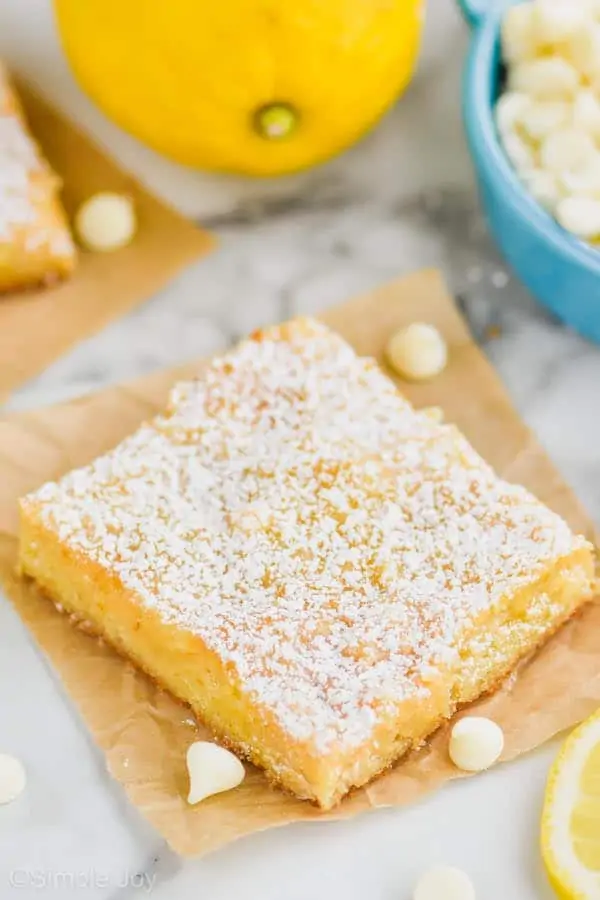 one lemon magic bar topped with powdered sugar on a square of parchment paper with white chocolate chips and lemons around