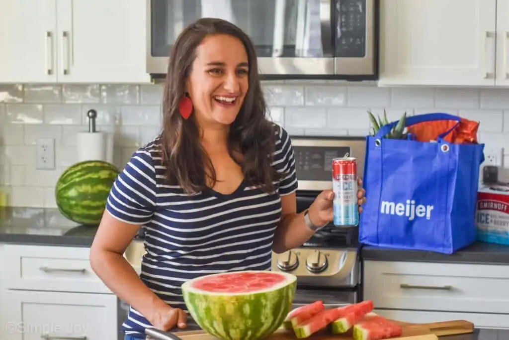 a woman holding a red white and blue can in a kitchen with a cut up watermelon and a reusable Meijer grocery bag in the background