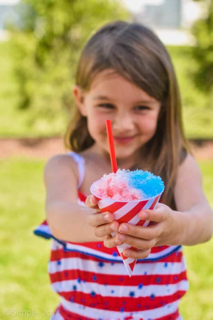 close up of a red and blue snow cone in a paper liner held by a little girl in a red and white striped dress