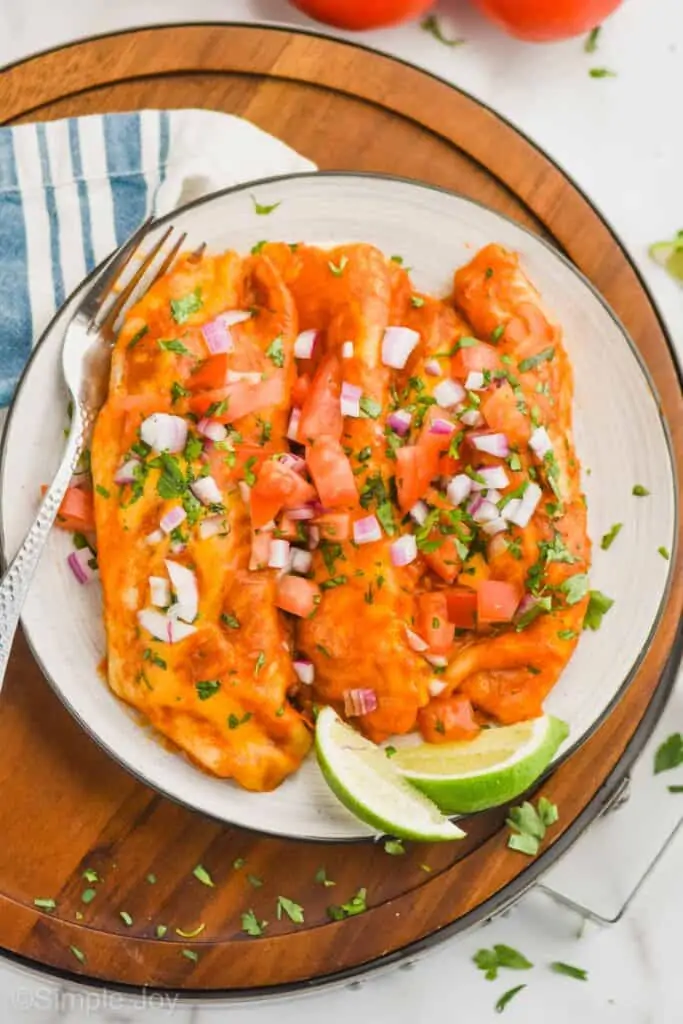 overhead view of a plate holding three crock pot chicken enchiladas garnished with cut up cilantro, red onion, tomatoes, and two lime wedges on the side