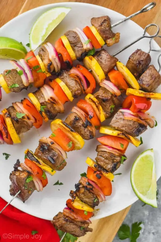 four steak kabobs on a white round plate with lime wedges around them