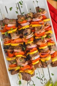 a pile of steak kabobs made with red peppers, yellow zucchini, and red onions with metal skewers on a white plate and garnished with parsley