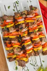 a pile of steak kabobs made with red peppers, yellow zucchini, and red onions with metal skewers on a white plate and garnished with parsley