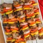 a pile of steak kabobs made with red peppers, yellow zucchini, and red onions with metal skewers on a white plate and garnished with parsley