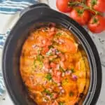 overhead view of a slow cooker full of crock pot chicken enchiladas garnished with cilantro, diced red onions and cut up tomatoes