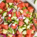 overhead view of a watermelon salad with mint, cucumbers, red onions, and feta in a white bowl with silver serving spoon and fork
