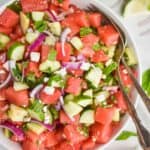 overhead view of a watermelon salad with mint, cucumbers, red onions, and feta in a white bowl with silver serving spoon and fork - 5