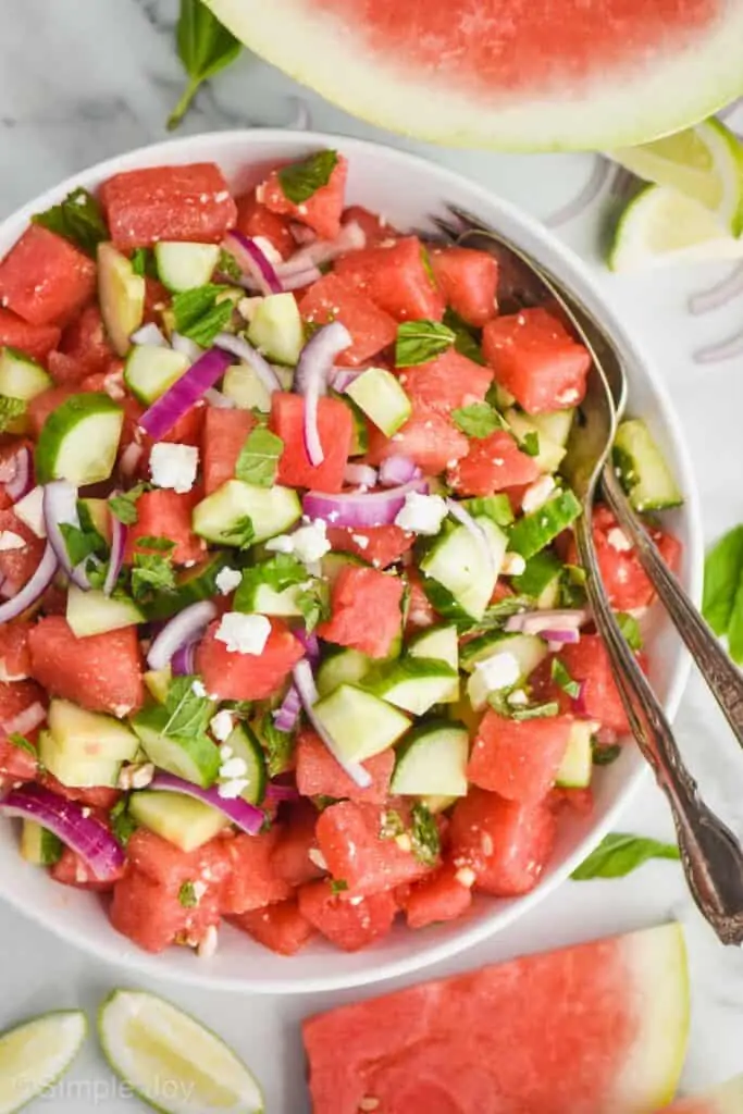 overhead view of a watermelon salad with mint, cucumbers, red onions, and feta in a white bowl with silver serving spoon and fork