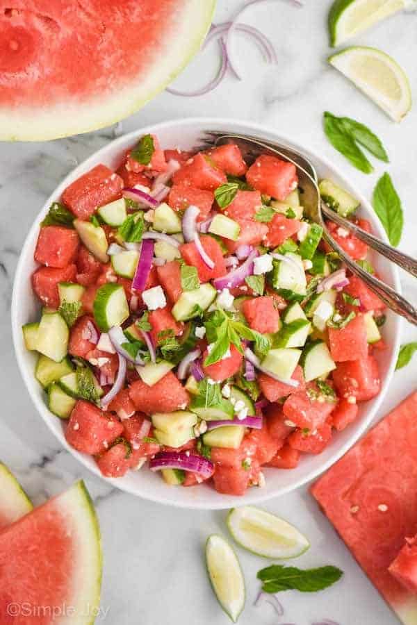 overhead view of a white bowl with watermelon salad in it, full pieces of watermelon, lime wedges, and mint leaves around on the white countertop