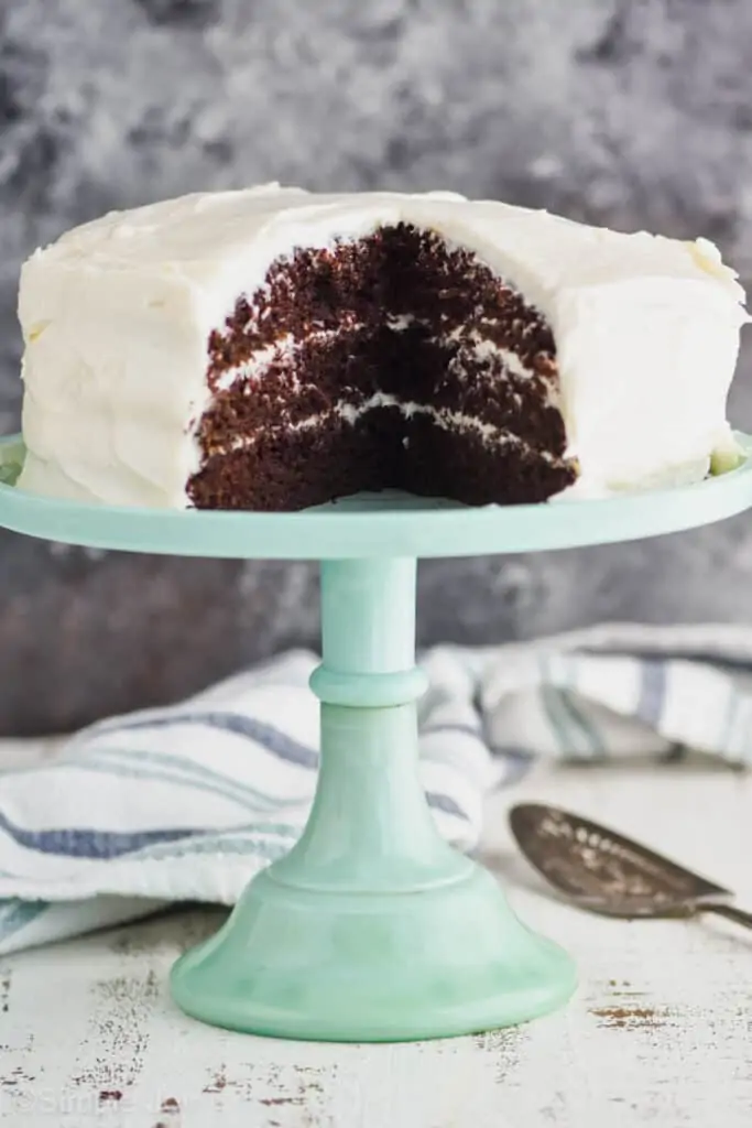 a three layer chocolate zucchini cake recipe on a teal cake stand with cream cheese frosting that has been cut into