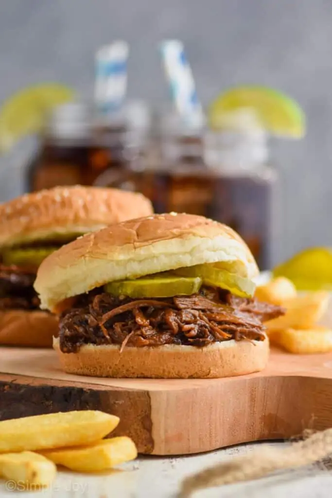 a bbq beef sandwich with pickles on it on a cutting board, fries in the foreground, cokes in the background