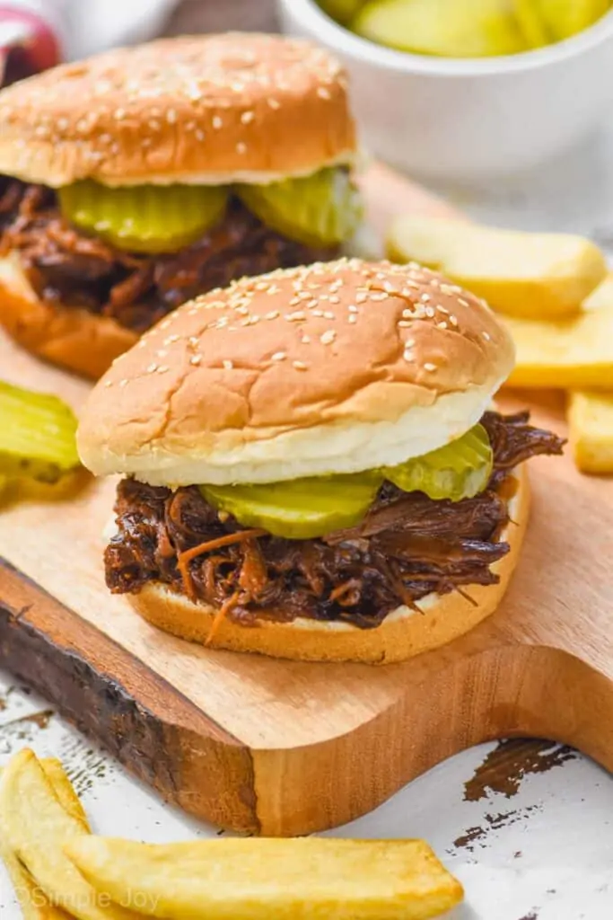 two crockpot bbq beef sandwiches on a cutting board, surrounded fries