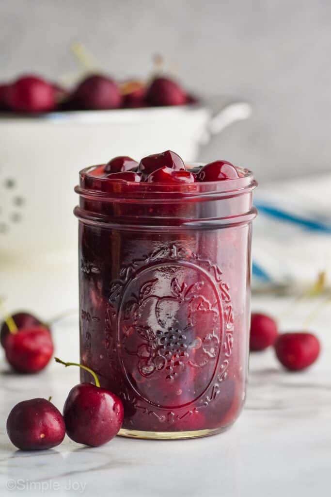 mason jar full of cherry pie filling with colander full of fresh cherries in the background