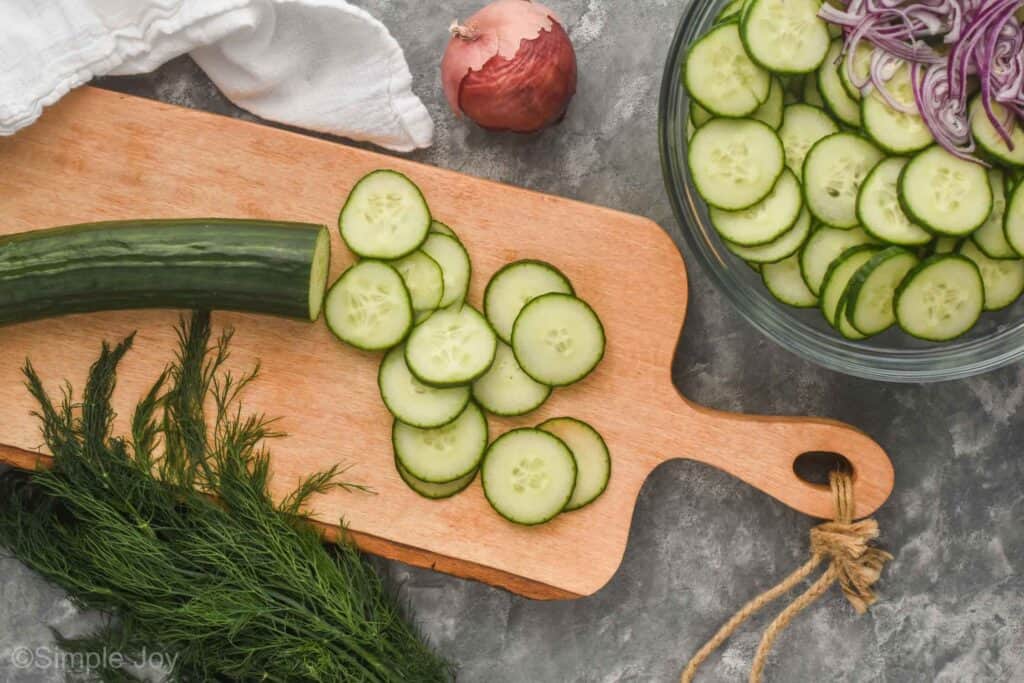 overhead view of a cucumber thinly slices on a cutting board next to fresh dill and a red onion