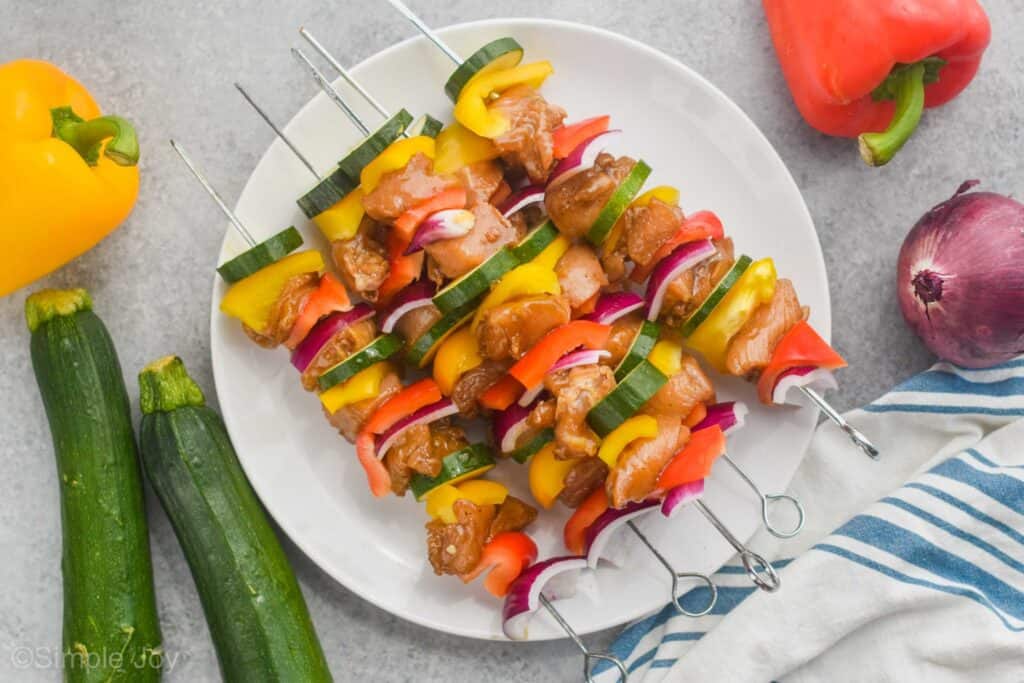 raw chicken kabobs on a white plate with metal skewers