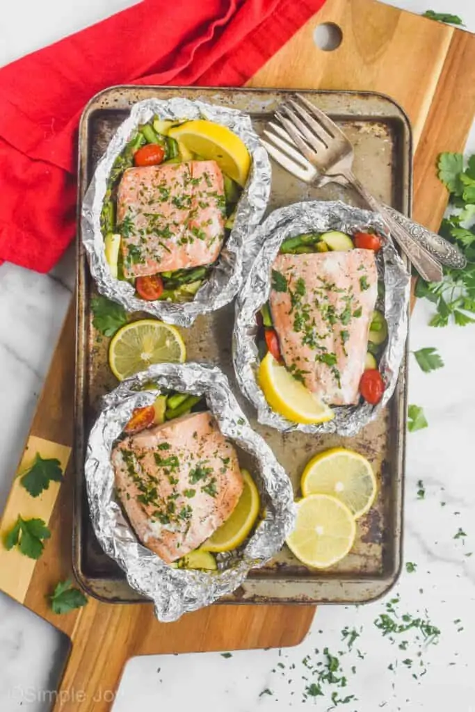 three salmon fillets in foil packets that were cooked with vegetables and garnished with parsley with fresh lemons around