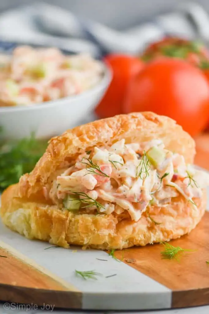 croissant sandwich with seafood salad on it, garnished with fresh dill, tomatoes in the background