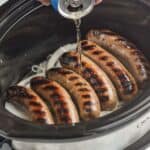 beer being poured over grilled brats with onions underneath in a layer in a slow cooker